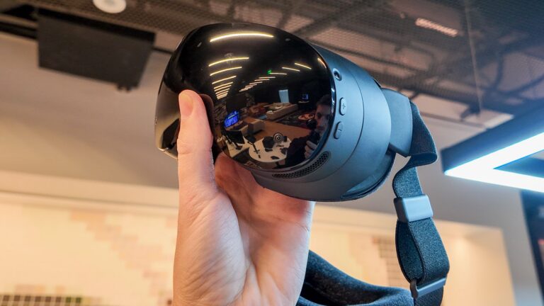 Eine Hand haelt das VR-Headset Steam Frame von Valve mit glaenzender schwarzer Frontblende und Stoff-Kopfband