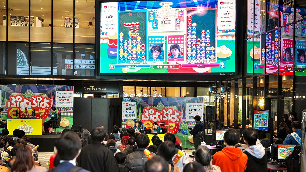 Öffentliches Puyo-Puyo-Champions-Esports-Event auf einer Großleinwand in Japan, davor sitzt eine Zuschauermenge