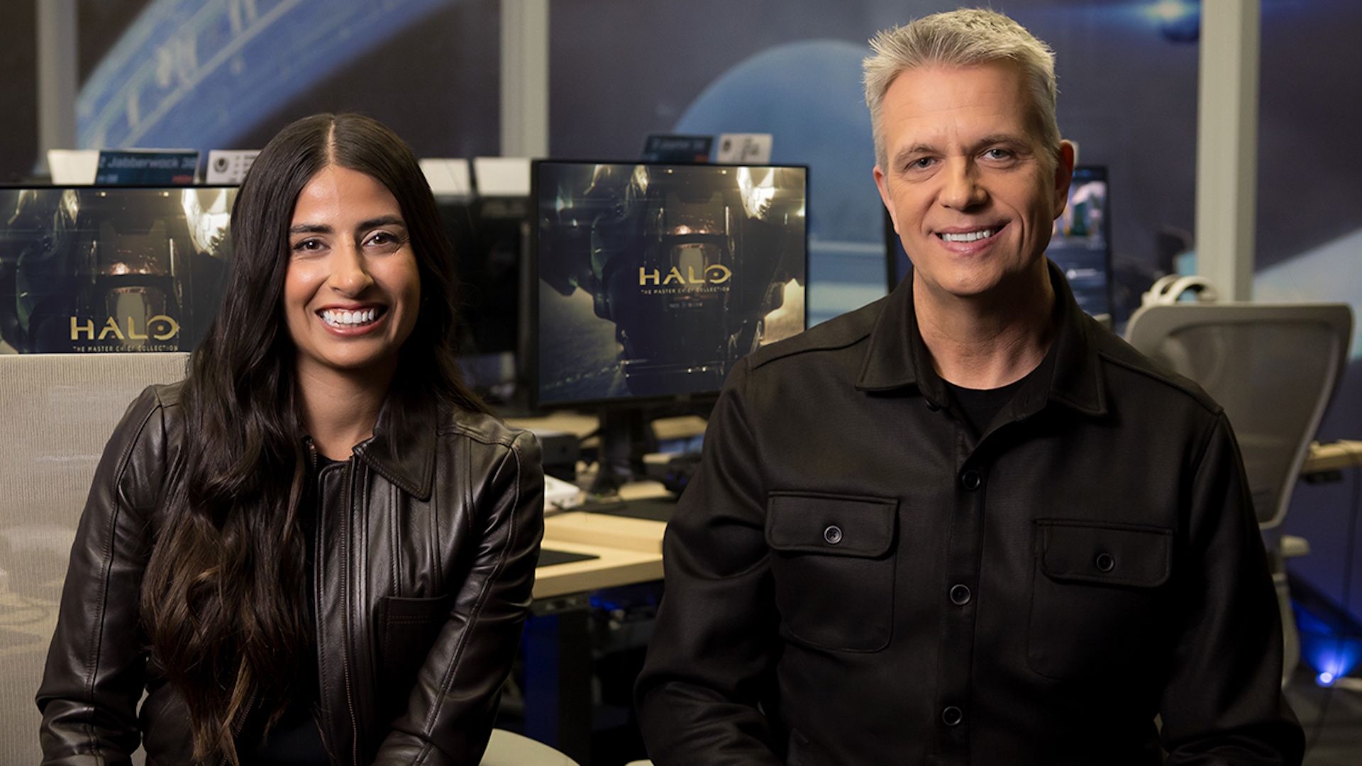 Zwei Mitarbeitende von Halo Studios sitzen lächelnd vor Bildschirmen mit Halo: The Master Chief Collection im Hintergrund
